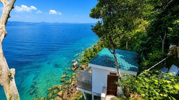 Una propiedad costera con vista despejada al mar, un pequeño edificio con balcón y abundante vegetación.