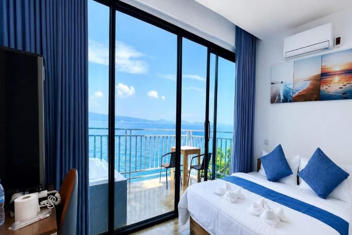 A hotel room with a large bed, a desk with a chair, a balcony with a view of the sea, and a painting on the wall.