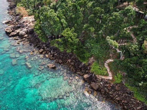 Vue aérienne d’une zone côtière avec des eaux turquoise limpides, une côte rocheuse, une végétation verdoyante et luxuriante, et un sentier sinueux.