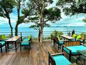 Una terraza exterior con suelo de madera, asientos acolchados de color verde y vista al mar.