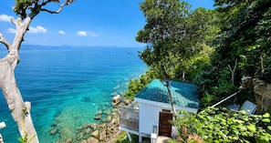 A coastal property with a clear view of the sea, a white building with a green roof, and lush greenery.