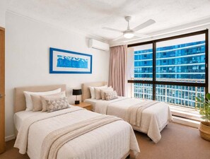 A hotel room with two beds, a ceiling fan, a large window with a view of a building, and a framed picture on the wall. - Beachfront Ocean Views | Pools, Spa & Tennis (Gold Coast)