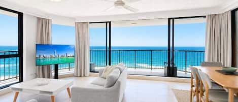 A modern living room with a sofa, a dining table, and a TV. The room has large windows with a view of the ocean.