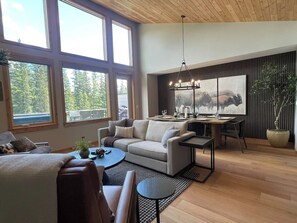 A modern living room with a sofa, coffee table, dining table, and a large window with a view of trees.