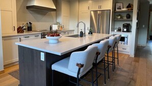 A modern kitchen with a central island, bar stools, stainless steel appliances, and a herringbone-patterned backsplash.
