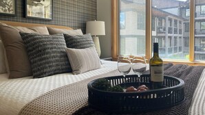 Une chambre à coucher avec un lit, des oreillers, une table de chevet avec une lampe, une fenêtre donnant sur des immeubles, et un plateau avec des verres à vin et une bouteille.