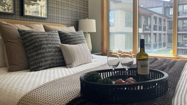 A bedroom with a bed, pillows, a bedside table with a lamp, a window with a view of buildings, and a tray with wine glasses and a bottle.