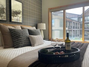 A bedroom with a bed, pillows, a bedside table with a lamp, a window with a view of buildings, and a tray with wine glasses and a bottle.