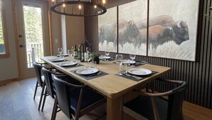A dining area with a long table set for six, wooden chairs, a chandelier, and a large wall-mounted painting of bison.