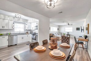 Une cuisine moderne avec des meubles blancs, un coin repas avec une table en bois dressée pour quatre personnes, et un salon avec un canapé et une table basse.