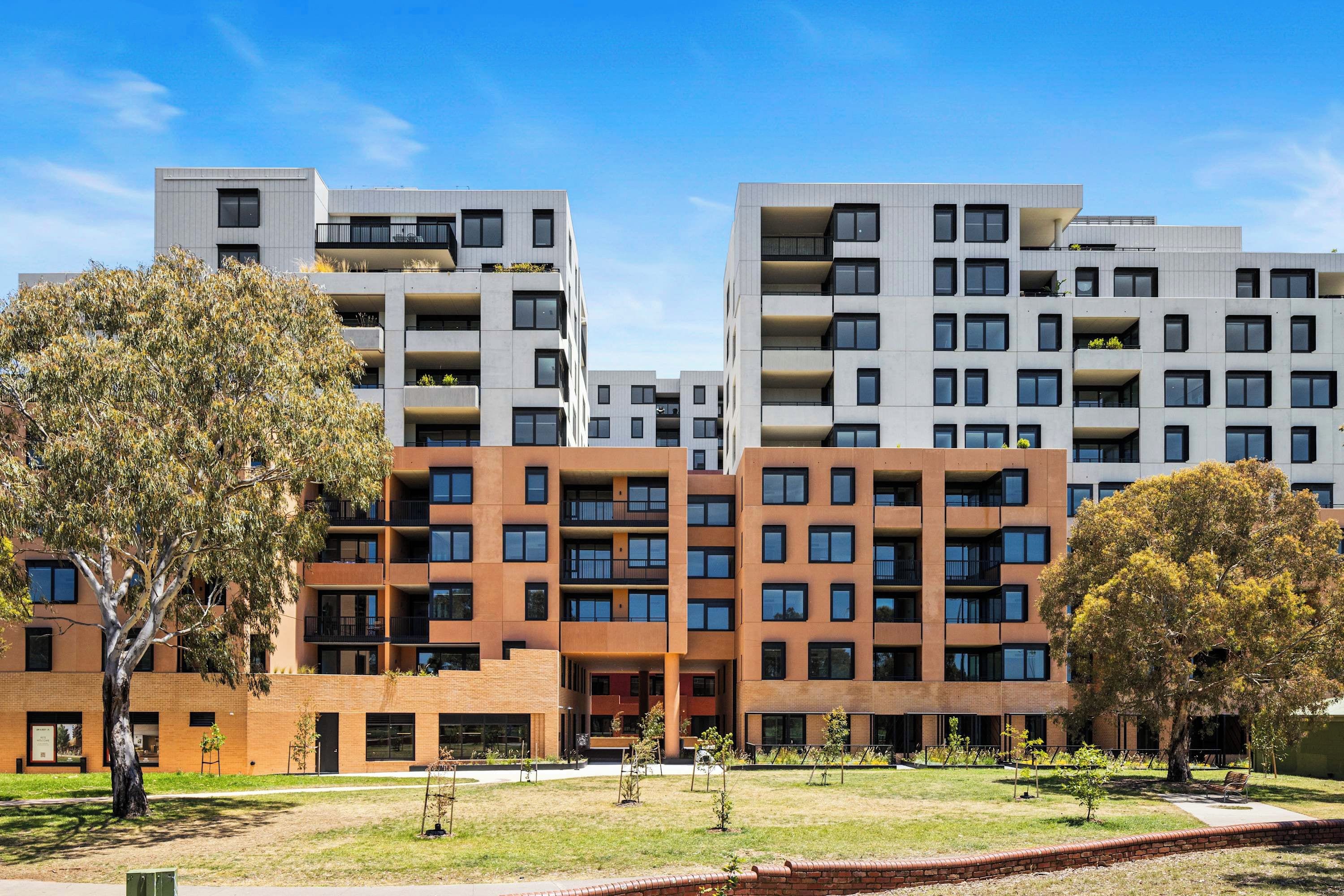 A modern multi-story residential building with balconies, a grassy courtyard, and trees.