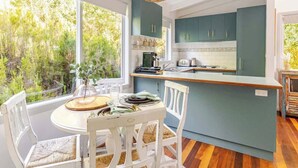 A kitchen with a dining area, large windows, and a view of greenery. - Cosy 2 Bedroom Cottage with spabath and fireplace (Kalorama)