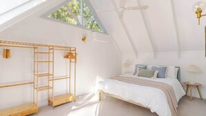 A bedroom with a sloped ceiling, a bed with a white and beige bedspread, a wooden bench, a wall-mounted shelf, and a ceiling fan. - Cosy 2 Bedroom Cottage with spabath and fireplace (Kalorama)