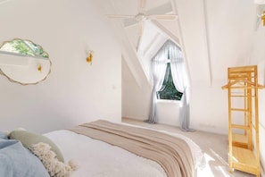 Ein Schlafzimmer mit einem Bett, einem Deckenventilator, einem Fenster mit Vorhängen und einer Holzleiter.