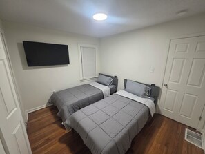 Ein Schlafzimmer mit zwei Betten, einem an der Wand befestigten Fernseher, einem Fenster mit Jalousien und einer Tür.