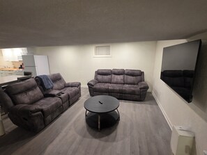 Ein Wohnzimmer mit zwei Liegesofas, einem Couchtisch, einer Küche im Hintergrund und einem Wand-Fernseher.