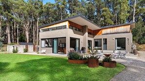 Una casa moderna de dos pisos con un amplio jardín delantero, un área de estar al aire libre y vistas a árboles altos.