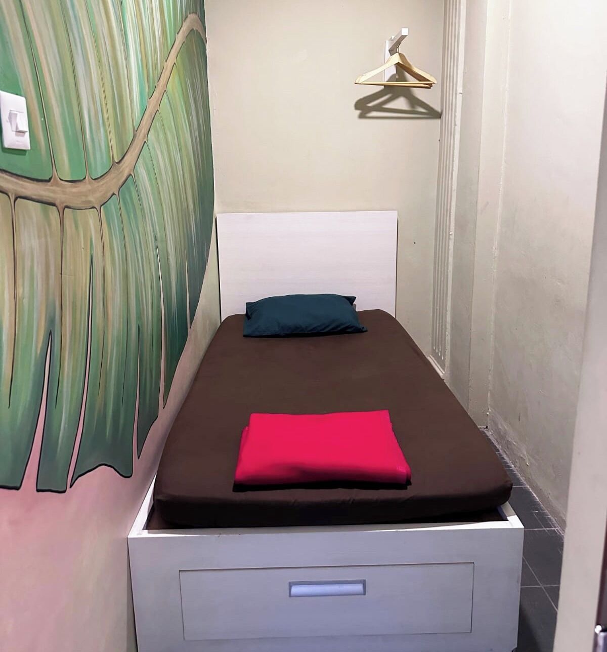 A single bed with a brown mattress and a red blanket, a white headboard, a blue pillow, a wall-mounted light switch, and a wooden hanger on the wall.