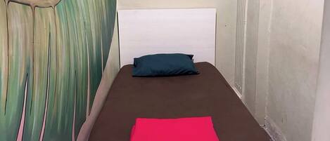 A single bed with a brown mattress and a red blanket, a white headboard, a blue pillow, a wall-mounted light switch, and a wooden hanger on the wall.