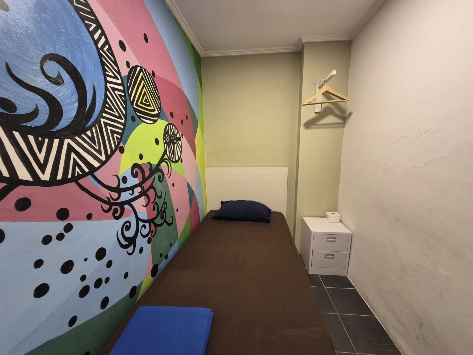 Una stanza piccola e luminosa, con un letto, una panca, un muro decorato con un murale colorato, un tavolino con una scatola e un appendiabiti.