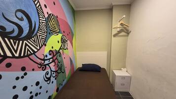 Una stanza piccola e luminosa, con un letto, una panca, un muro decorato con un murale colorato, un tavolino con una scatola e un appendiabiti.