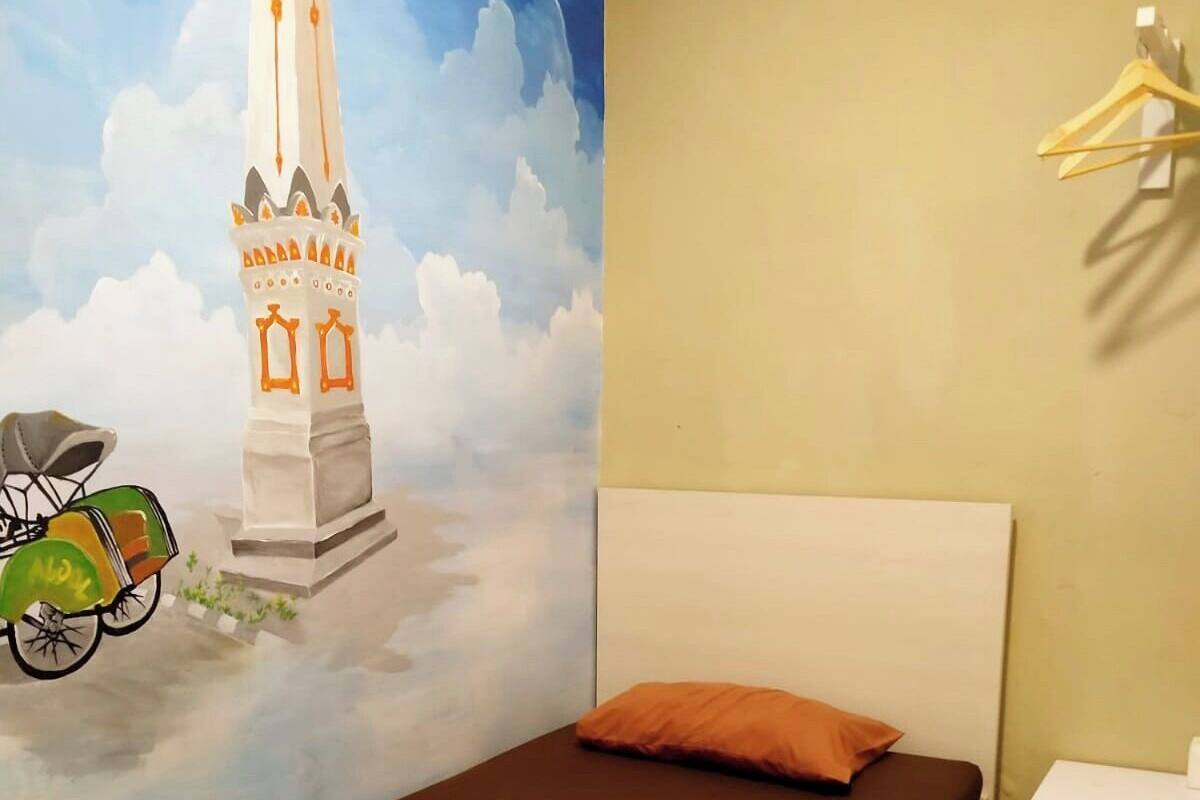 A bed with a brown mattress and a single orange pillow, a white headboard, a wall with a mural, a small bedside table, and a hanging light fixture.