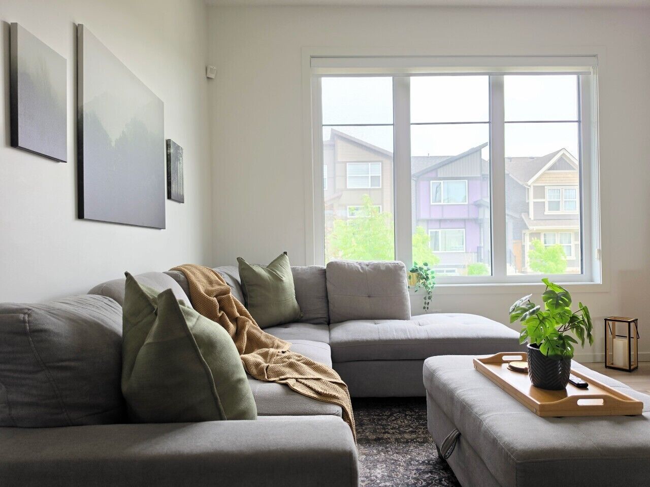 Ein Wohnzimmer mit einer Eckcouch, einem Couchtisch mit Pflanze und einem Fenster mit Blick auf Häuser.