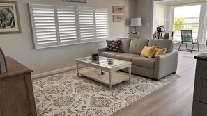 Ein Wohnzimmer mit einer Couch, einem Glastisch, einem Teppich mit Muster und einem Fenster mit Jalousien.