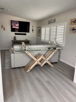 A modern living room with a glass coffee table, a sofa, a TV mounted on the wall, and a window with blinds. - Rest Ashore on the Sarasota Bay (Bradenton)