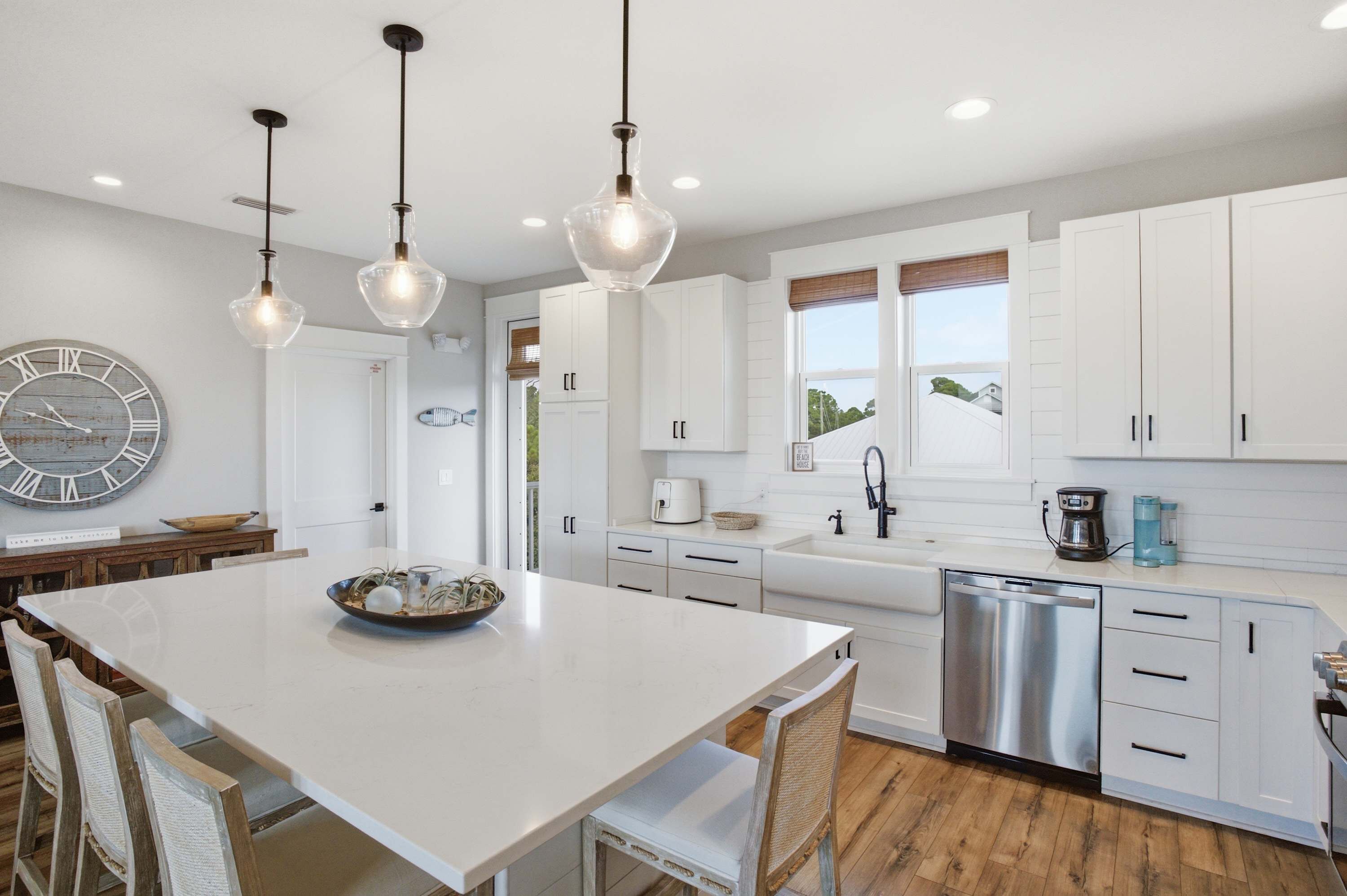 A modern kitchen with white cabinetry, a central island with seating, pendant lighting, and a large wall clock.