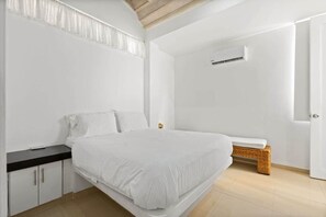 Quarto com cama, mesa de cabeceira, pequeno banco, porta e aparelho de ar condicionado.
