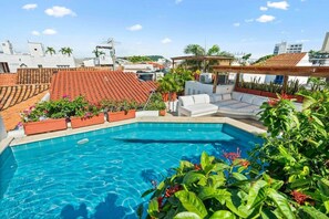 Piscina no terraço com água azul cristalina, mobiliário de exterior branco e telhados vermelhos ao fundo.