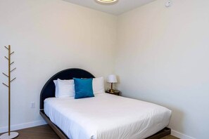 A neatly made bed with white and blue pillows, a dark headboard, a bedside lamp, and a coat rack. - The Gallery | Lux Studio in Wynwood (Miami)