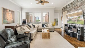 A modern living room with a sectional sofa, a leather couch, a coffee table, a ceiling fan, and a large TV on a brick wall. - Sky house in LasColinas near Dallas Stadium in Arlington (Irving)
