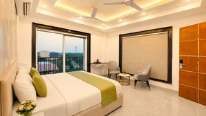 Quarto de hotel moderno com uma cama grande, duas cadeiras, uma mesinha e vista para a cidade.