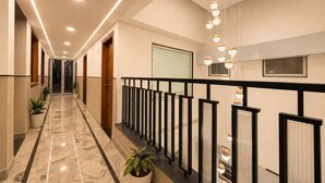 Corredor de hotel com piso de mármore, plantas em vasos, corrimão preto e iluminação pendente.