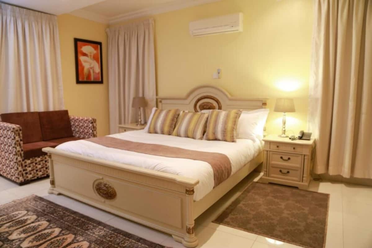 A hotel room with a bed, a nightstand, a chair, a rug, and an air conditioning unit.