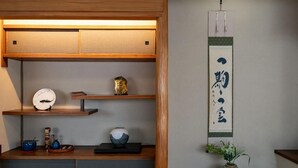 Ett traditionellt japanskt rum med tatamimattor på golvet, en låg trähylla med dekorativa föremål, en upphängd rullbild med kalligrafi och en liten växt på en tatamimatta.