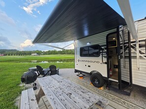 A travel trailer with a covered patio area, a picnic table, and a view of a green field.