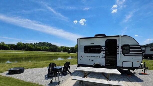 Una caravana blanca con la marca 'Campbound' estacionada junto a un lago, con mesas y sillas de picnic.