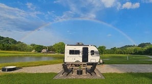 A white RV with a 'Campbound' logo parked on a gravel path with a picnic setup, a lake, and a rainbow in the background.