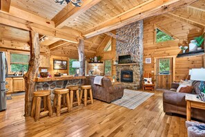 A log cabin interior with a stone fireplace, wooden bar stools, a sofa, and a kitchen area. - LINDEN LODGE/7BR Near Old Man's Cave/Hot Tub & Game Room (Logan)