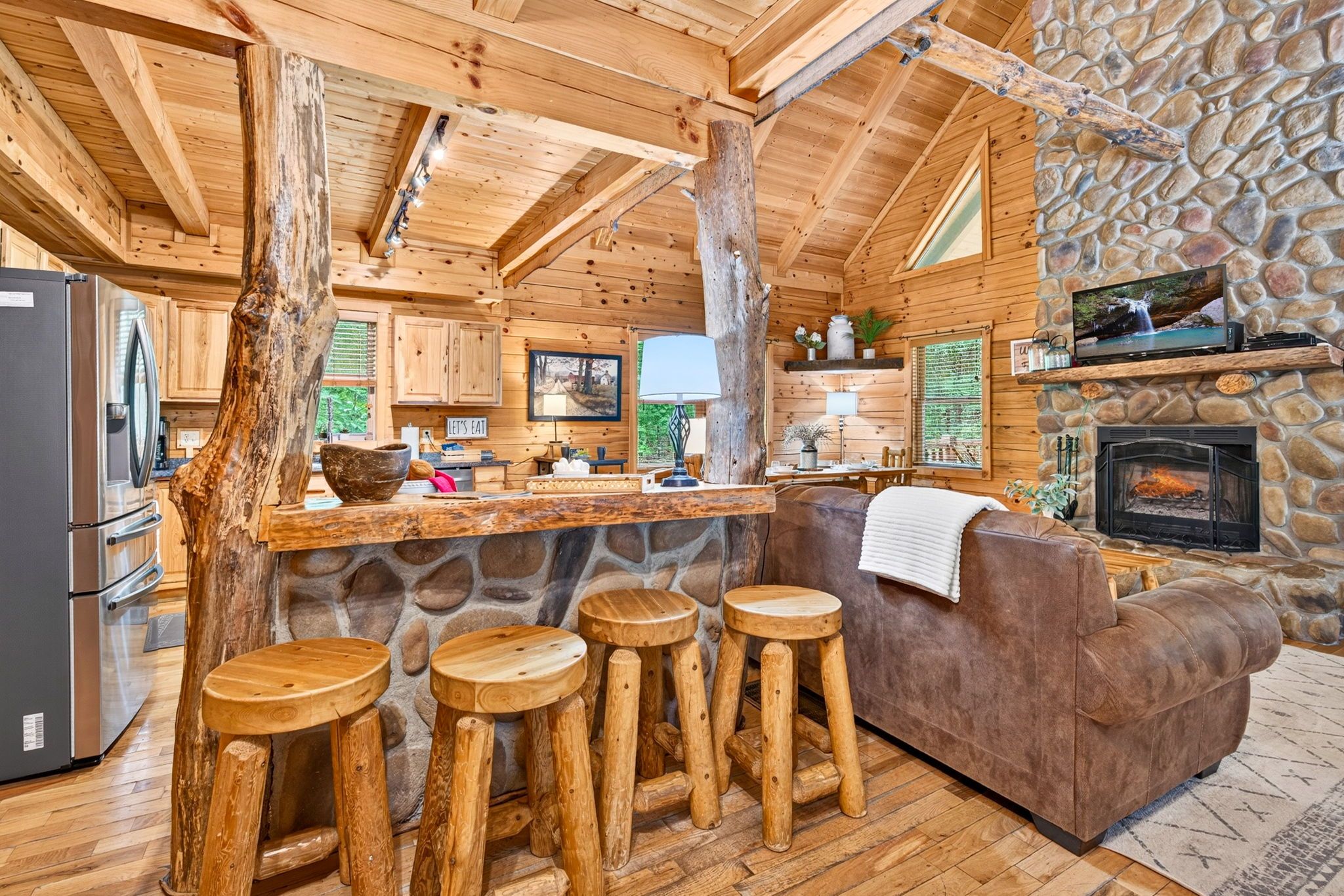 A rustic cabin interior with a stone fireplace, wooden bar stools, a sofa, and a kitchen area.