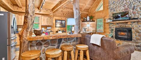 A rustic cabin interior with a stone fireplace, wooden bar stools, a sofa, and a kitchen area.