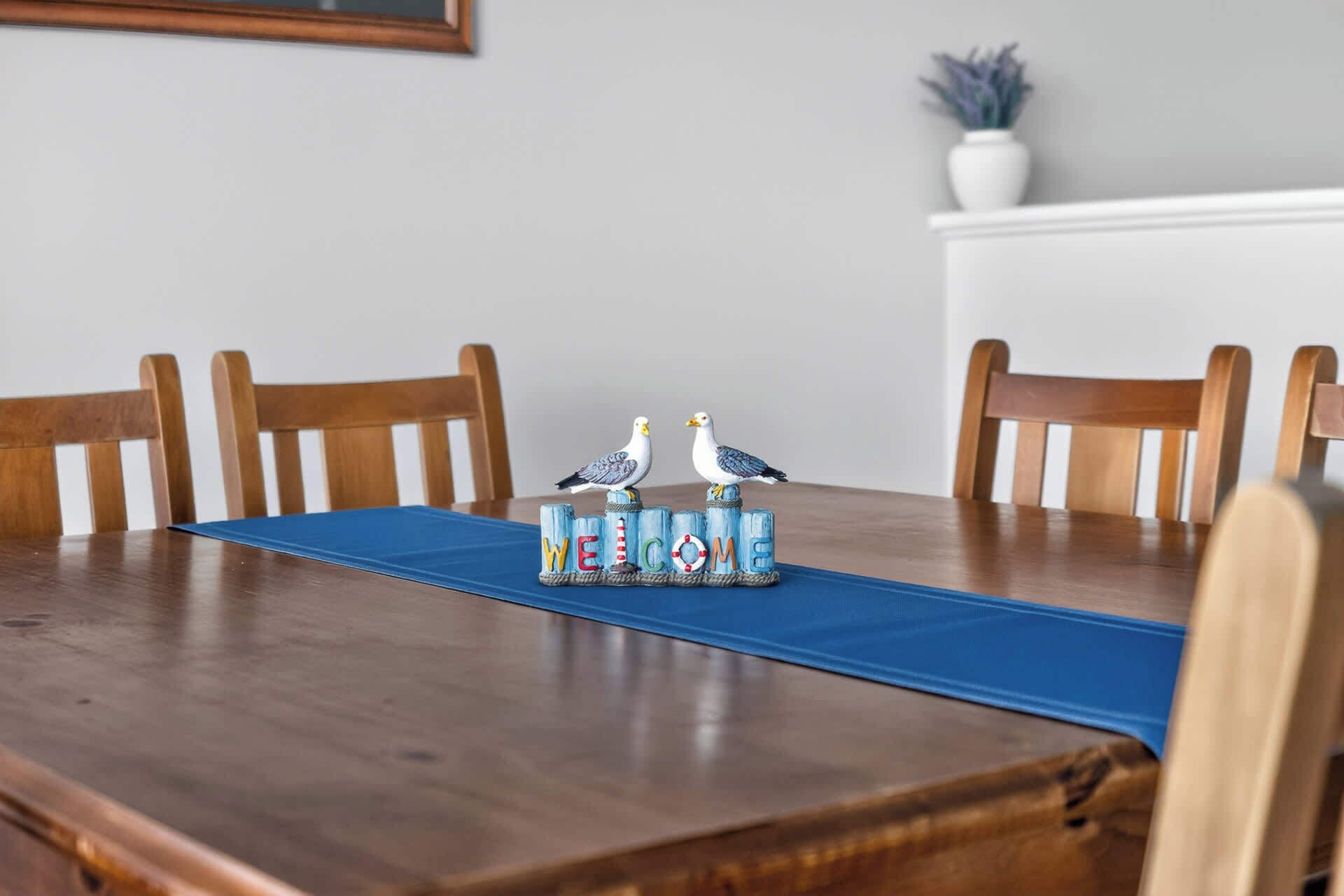 Une table de salle à manger en bois, surmontée d’un set de table bleu, accompagnée de deux chaises en bois et d’un panneau décoratif représentant un oiseau, sur lequel est inscrit le mot « BIENVENUE ».