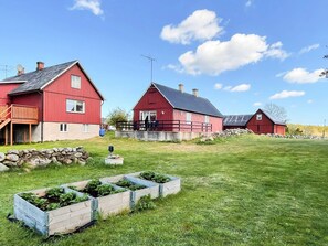 Ein rotes Bauernhaus mit Steinmauer, einer Holzterrasse und einem grasbewachsenen Feld mit erhöhten Blumenbeeten.