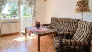 Une pièce avec un plancher en bois, un canapé à rayures, une table recouverte d’une nappe rouge, une lampe et une fenêtre ornée de rideaux à motifs floraux.