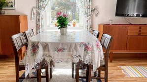 Un coin repas avec une table dressée pour six personnes, entourée de chaises, un téléviseur accroché au mur et une fenêtre avec des rideaux.