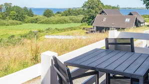Eine Holzterrasse mit Tisch und Stühlen mit Blick auf ein Gewässer, eine Wiese und ein Gebäude mit geneigtem Dach.