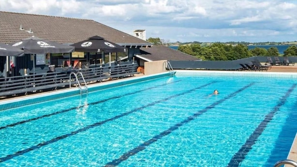 Ein Schwimmbecken mit klarem, blauem Wasser, ein Rettungsschwimmerstuhl und eine überdachte Sitzgelegenheit mit Sonnenschirmen.